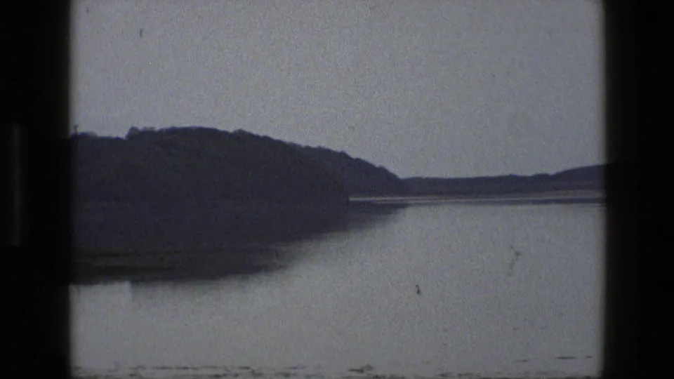 1969 England home-movie frame showing dark hills beyond still water under a grey sky.