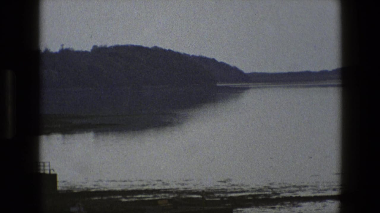 1969 England frame with still water, a dark hillside, muddy shore, and a small dock structure at the edge.