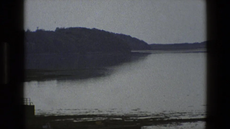 1969 England frame with still water, a dark hillside, muddy shore, and a small dock structure at the edge.