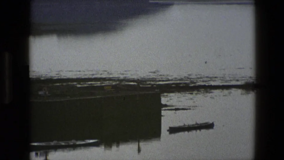 1969 England home-movie view of a swampy or muddy shoreline with a dock edge and small boat on calm water.