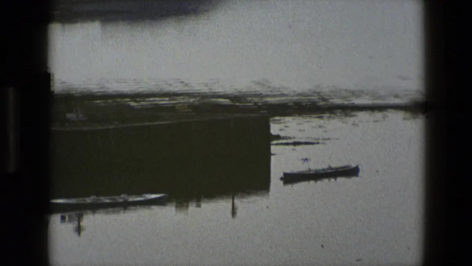 1969 England frame showing a small moored boat beside a dock or pier on a quiet, grey waterside scene.