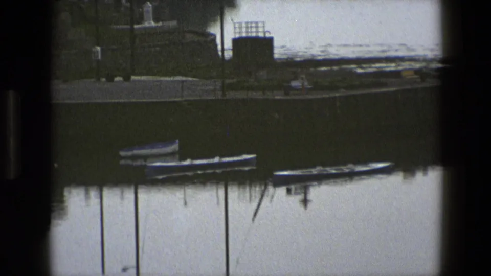 1969 England waterfront with moored small boats, a quay edge, posts, and distant structures reflected in calm water.