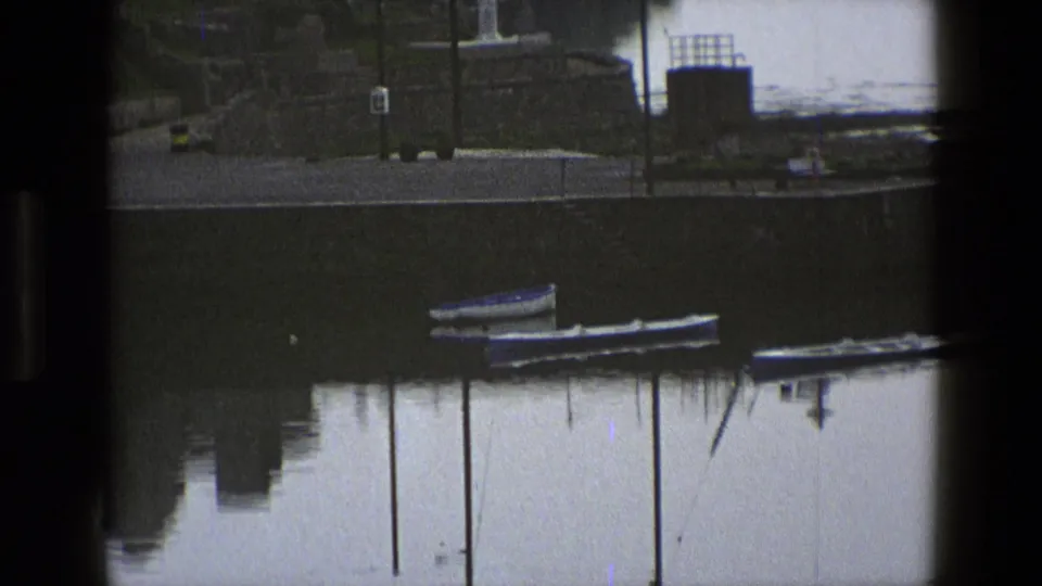 1969 England harbor frame showing small boats near mooring posts below a stone waterfront with tiny distant people visible.