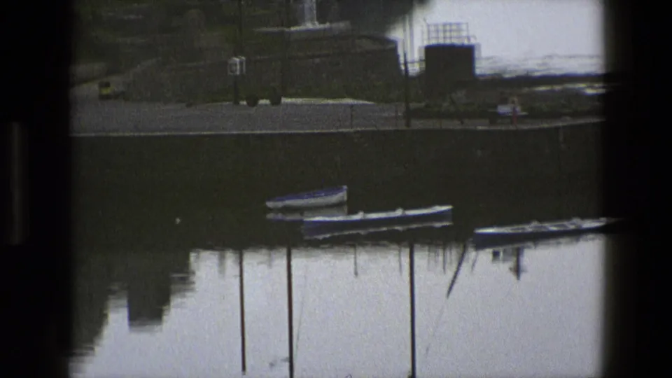 1969 England harbor view with boats, tall mooring posts, a stone quay, and a few tiny distant people near the waterfront.