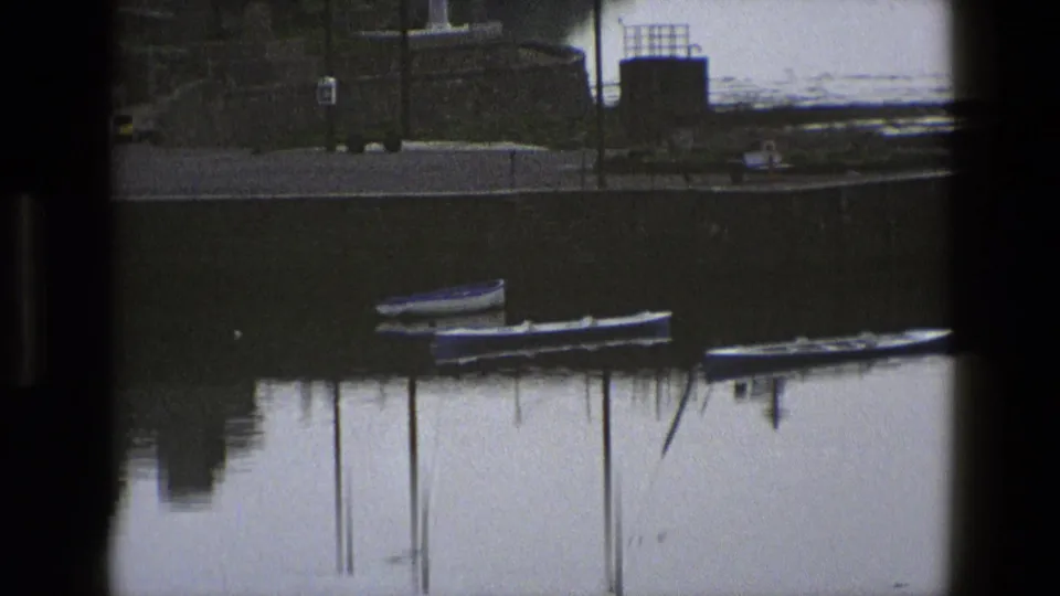 1969 England 8mm frame showing small moored boats and their reflections in still water under dark pier or window framing.