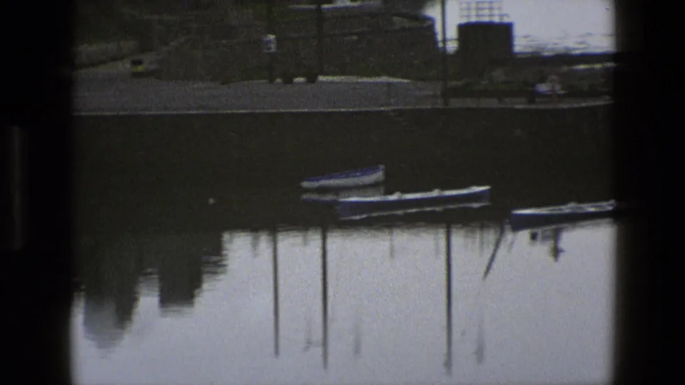 1969 England waterfront view with small boats, reflected posts, calm water, and dark foreground divisions from the film frame.