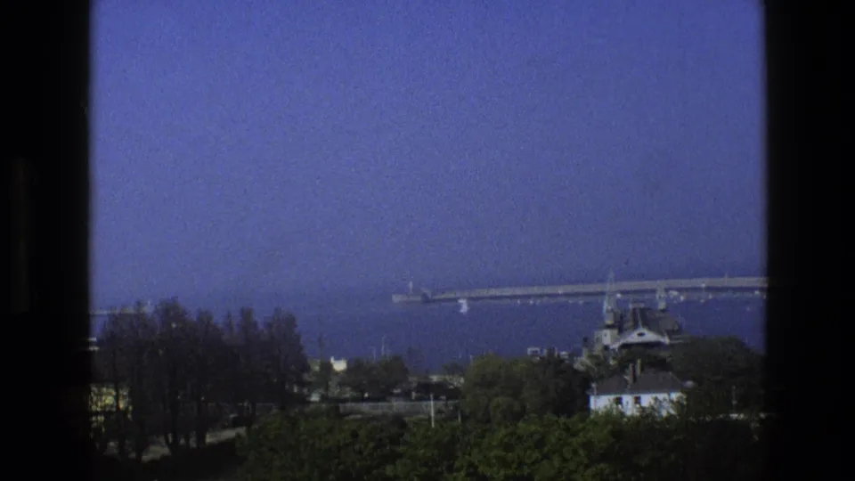 1969 England waterfront frame with a distant bridge, tree line, small building, open water, and blue color shift typical of aged 8mm film.