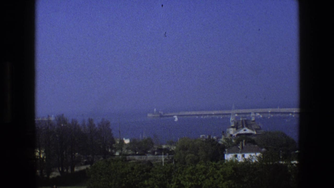 1969 England coastal view from behind dark window framing, with a long pier, calm sea, trees, and rooftops in the foreground.