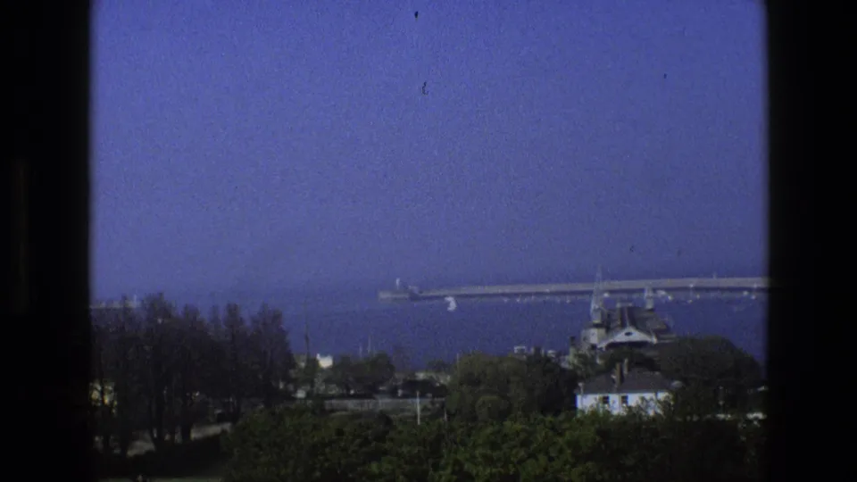 1969 England coastal view from behind dark window framing, with a long pier, calm sea, trees, and rooftops in the foreground.