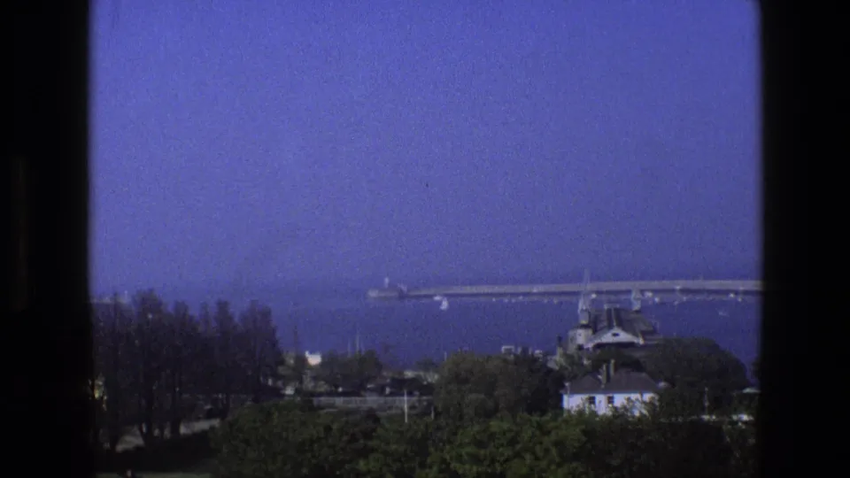 The 8mm frame holds on a blue-tinted coastal scene in England, with a long pier, trees, rooftops, and dark window borders.