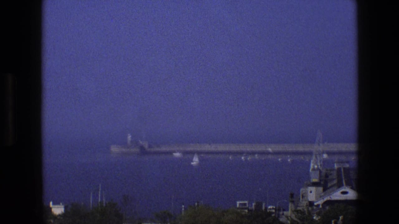 1969 England 8mm frame showing a calm bay with a long pier, small sailboats, shoreline trees, and dark window framing.