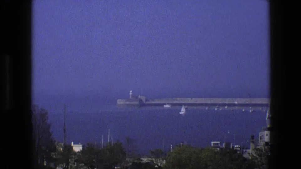 Grainy 1969 England 8mm still of a quiet bay with a pier, sailboats, treetops, rooftops, and dark window framing.