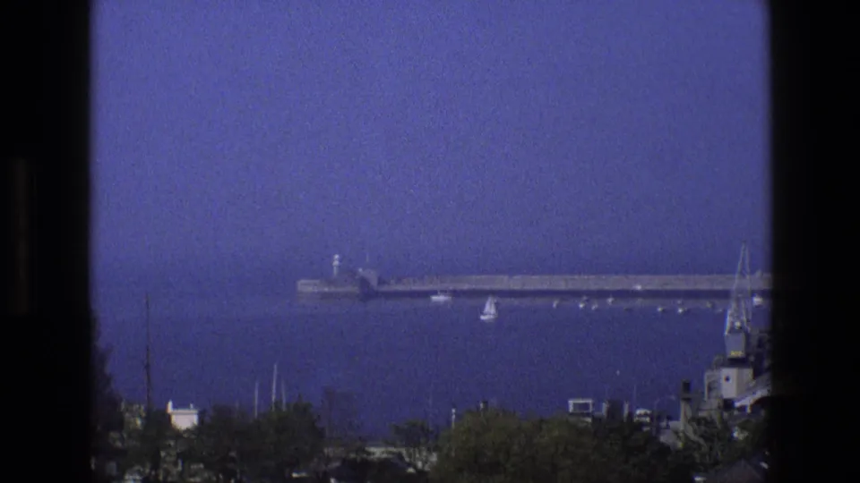 1969 England home-movie frame showing a distant pier, a small sailboat, calm bay water, and foreground shoreline trees.