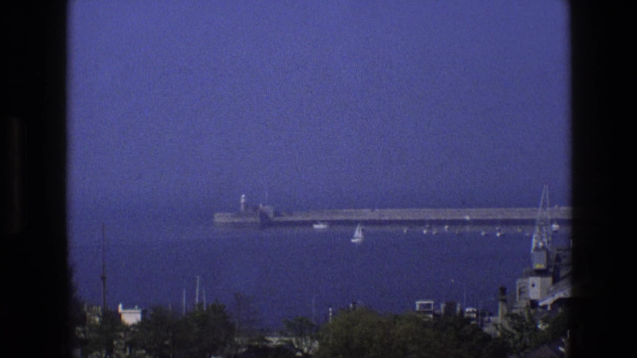 1969 England 8mm frame showing a hazy coastal harbor from above, with a long pier, small boats, shoreline greenery, and dark foreground framing.
