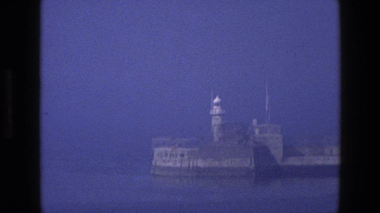 Zoomed harbor view of a stone breakwater with a small white lighthouse tower in haze.
