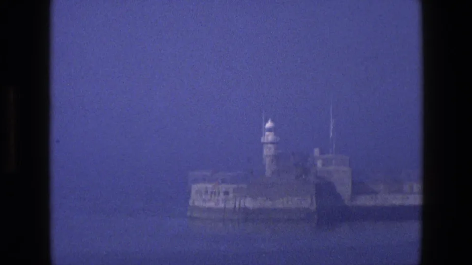 1969 England 8mm frame focused on a stone breakwater or fort-like harbor structure with a white lighthouse tower against hazy blue water and sky.