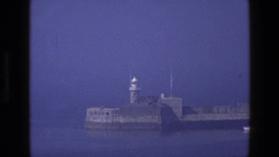 1969 England 8mm frame showing the lighthouse and stone harbor wall in soft focus, with calm blue water and a dark vertical edge at right.