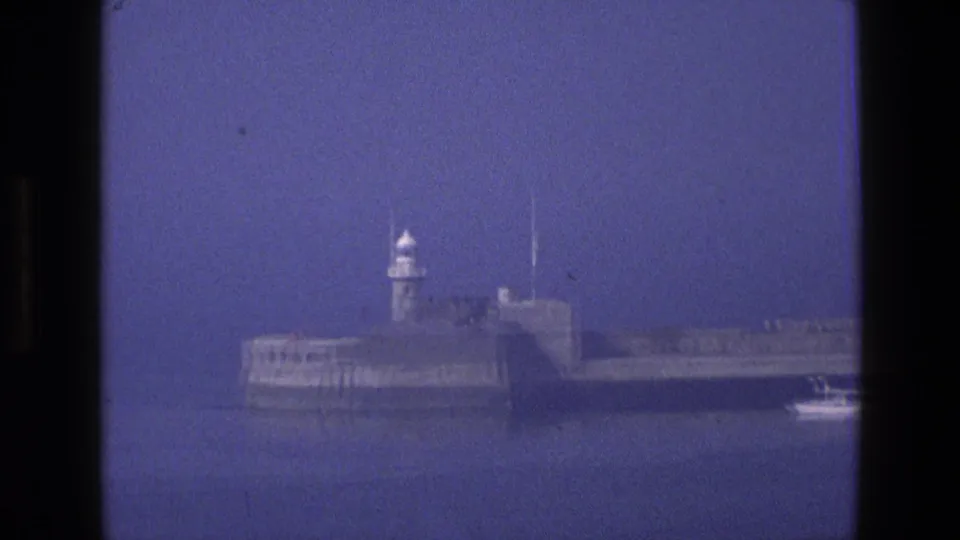 1969 England 8mm frame of the lighthouse breakwater with a small motorboat or vessel near the right edge, seen across blue harbor water.