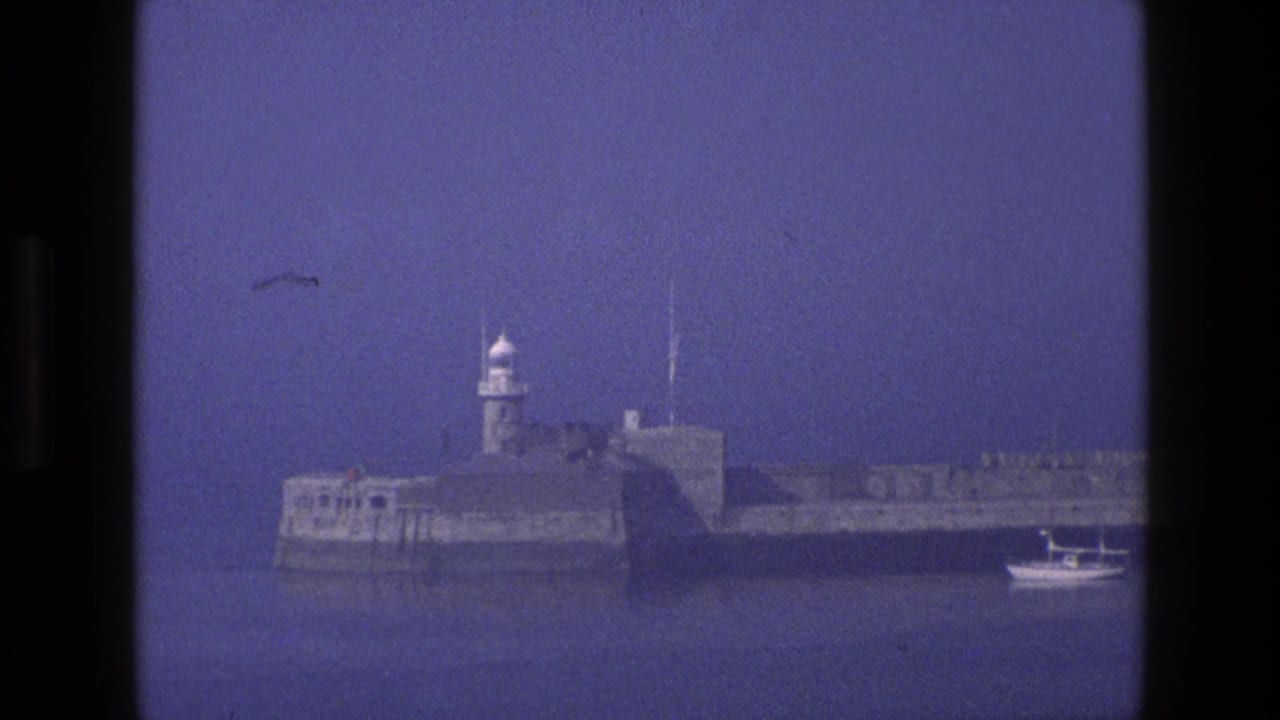 Stone coastal fort with a small lighthouse seen across calm blue water.