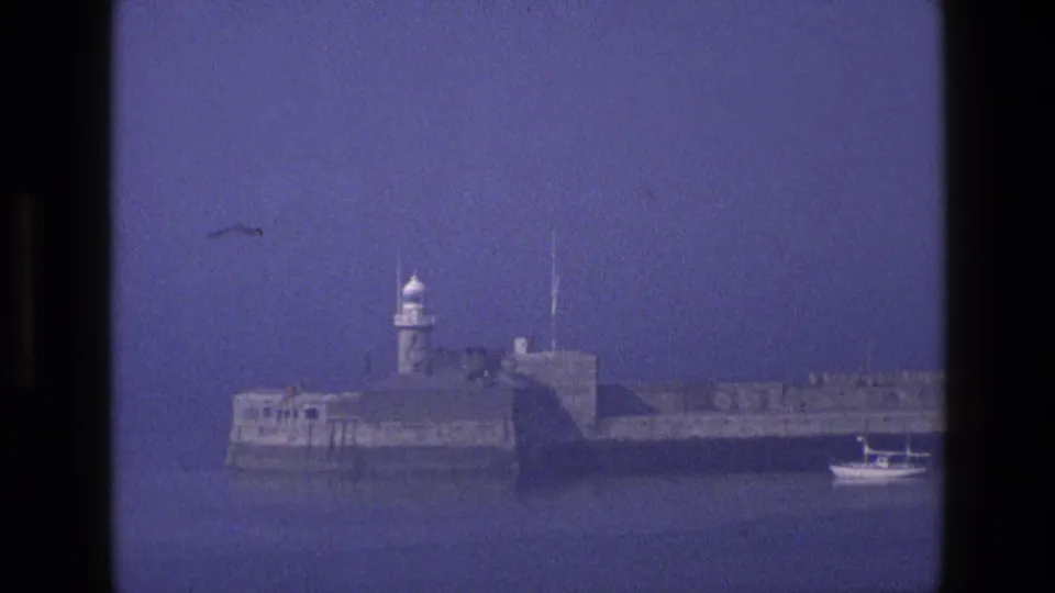 1969 England home-movie frame showing a stone harbor fort or breakwater with a small lighthouse and a boat at the right edge.