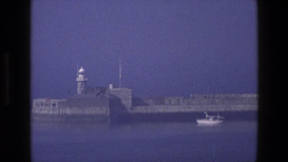 1969 England frame of a stone coastal structure with lighthouse, filmed from across the water as a small motorboat sits in the harbor.
