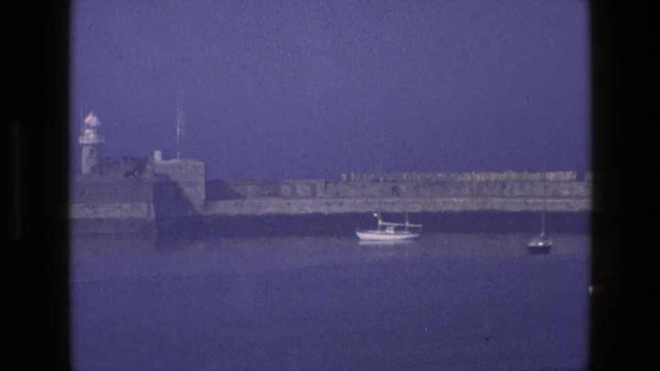 1969 England 8mm frame showing the fort and harbor wall with a motorboat and a small sailboat below the blue horizon.