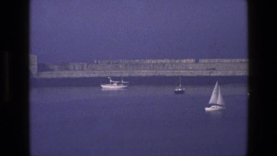 1969 England coastal home-movie frame with the harbor wall stretching across the background and small boats on blue water.