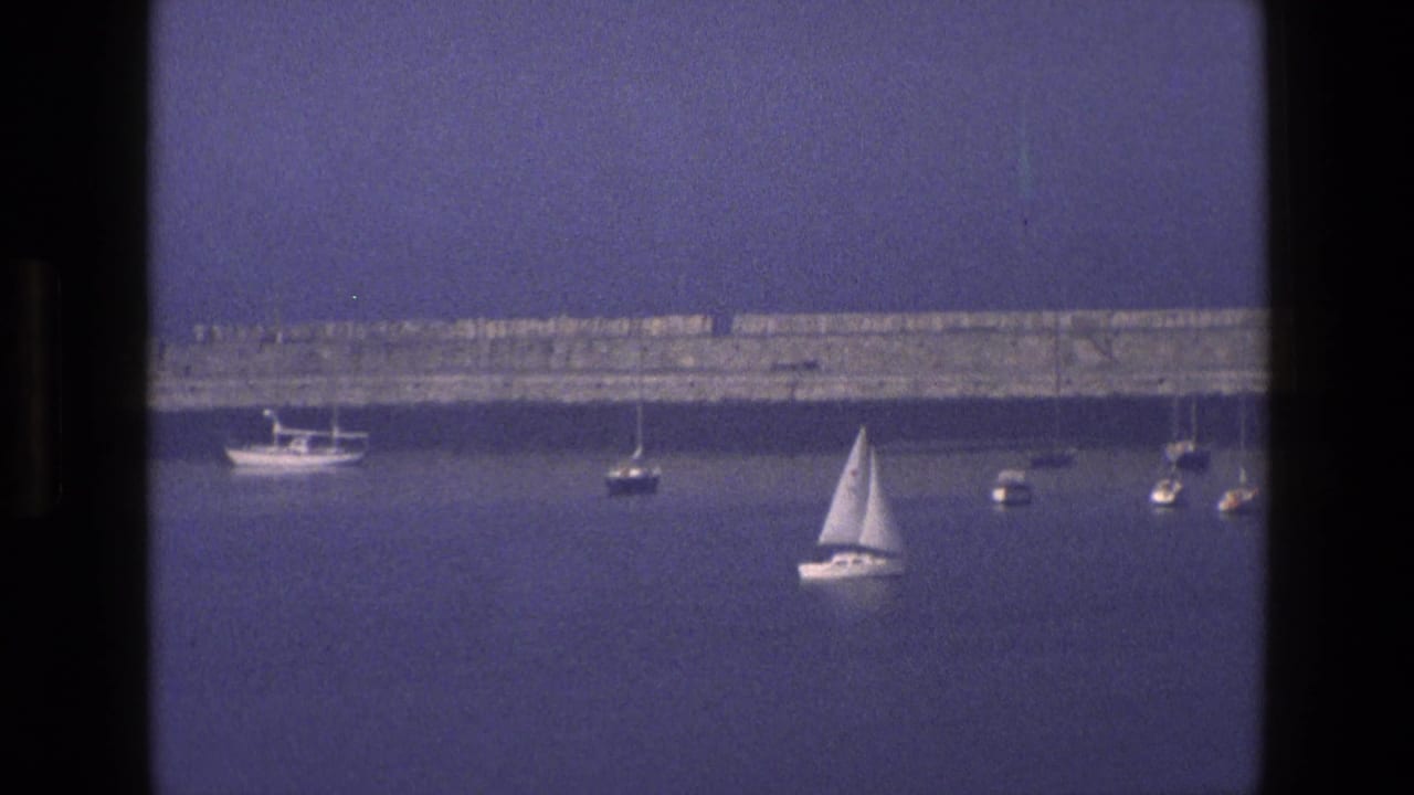 1969 England coastal home-movie frame with small boats and a sailboat on calm blue water, framed by dark window mullions.