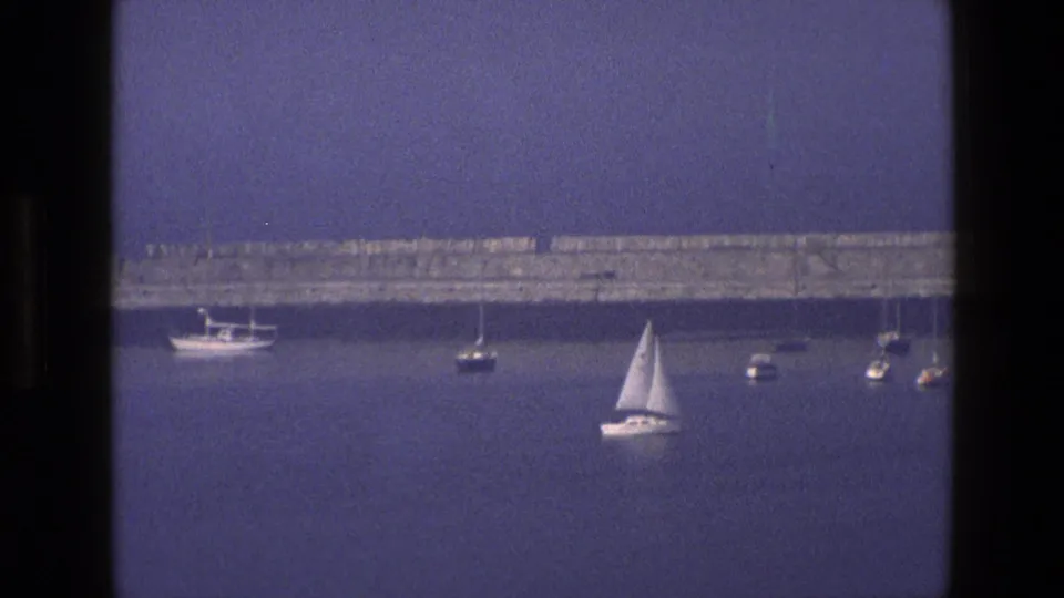 1969 England coastal home-movie frame with small boats and a sailboat on calm blue water, framed by dark window mullions.