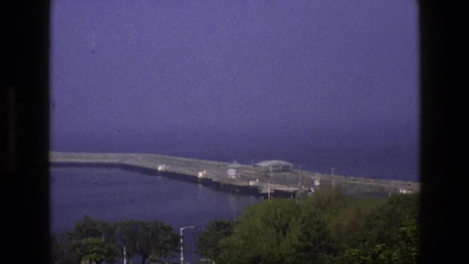 1969 England 8mm view of a long coastal pier or breakwater extending into blue water, with trees below the frame.