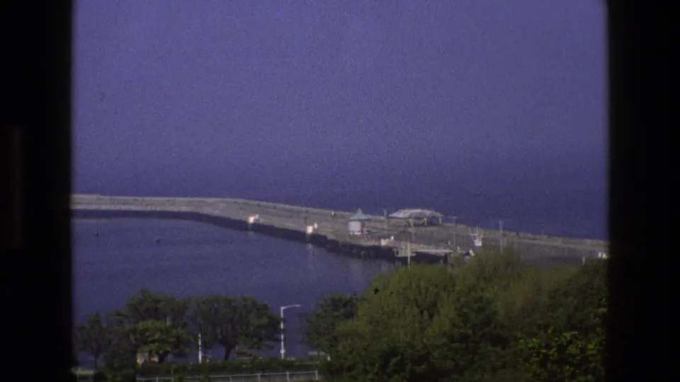 1969 England home-movie frame showing a stone pier beside calm bay water, with dark window framing and trees in the foreground.