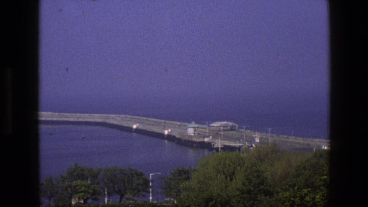 1969 England 8mm view of a long seaside breakwater, small shelters, trees, and calm water seen through a window frame.