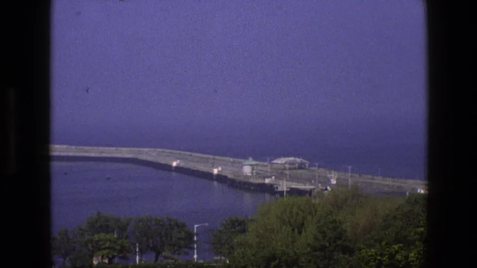 1969 England 8mm view of a long seaside breakwater, small shelters, trees, and calm water seen through a window frame.