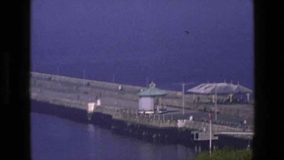 Closer 1969 England 8mm frame of a breakwater end with pavilion, shelter canopy, railings, water, and tiny distant visitors.