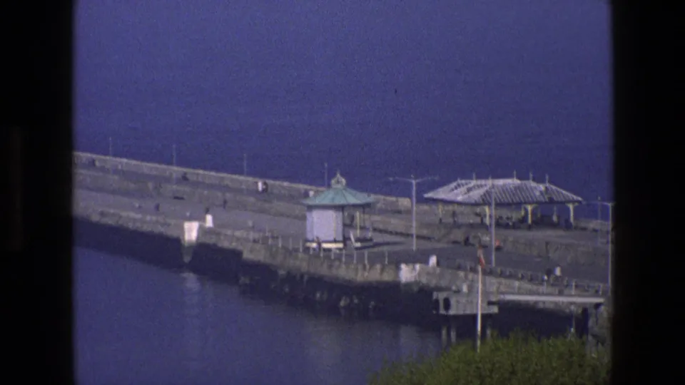 1969 England home-movie frame of the breakwater end, showing shelters, railings, calm water, foreground trees, and small visitors.