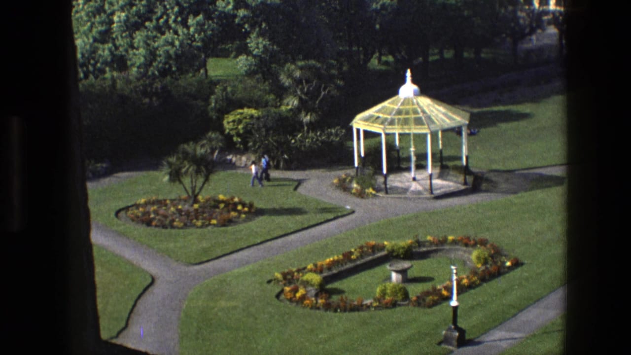 Elevated view of a formal garden with a white gazebo, flowerbeds, lawns, paths, and two walkers.