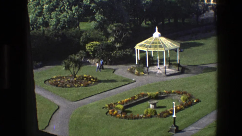 1969 England 8mm frame showing a formal garden from above, with a white gazebo, curving paths, flowerbeds, lawns, and distant visitors.