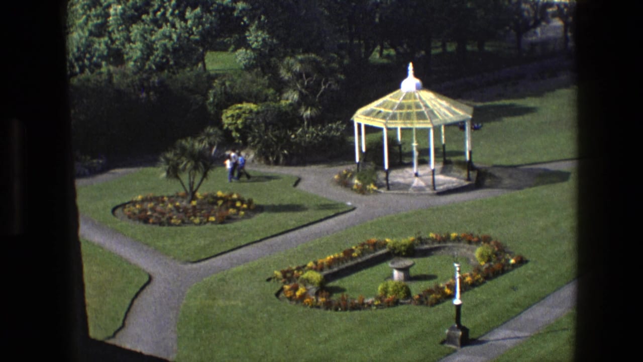 1969 England 8mm view of a formal garden, with visitors near shrubbery, a white gazebo, geometric paths, and planted beds.