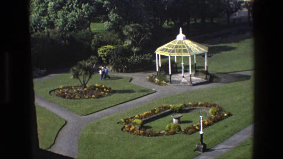 1969 England 8mm view of a formal garden, with visitors near shrubbery, a white gazebo, geometric paths, and planted beds.