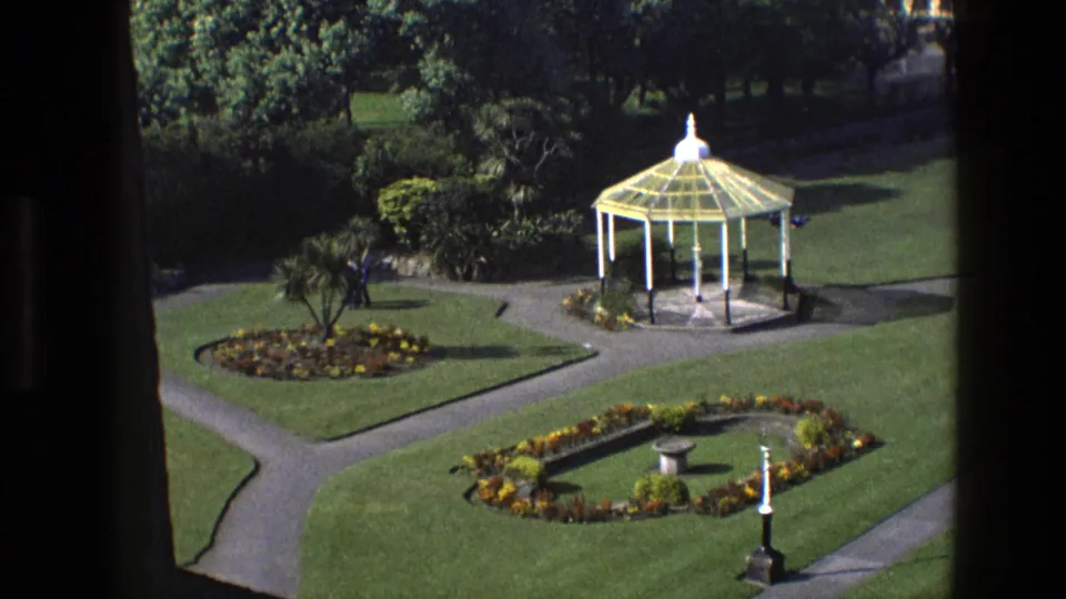 1969 England 8mm frame of a formal garden with a white gazebo, flowerbeds, a small central planter or fountain, and a distant visitor.