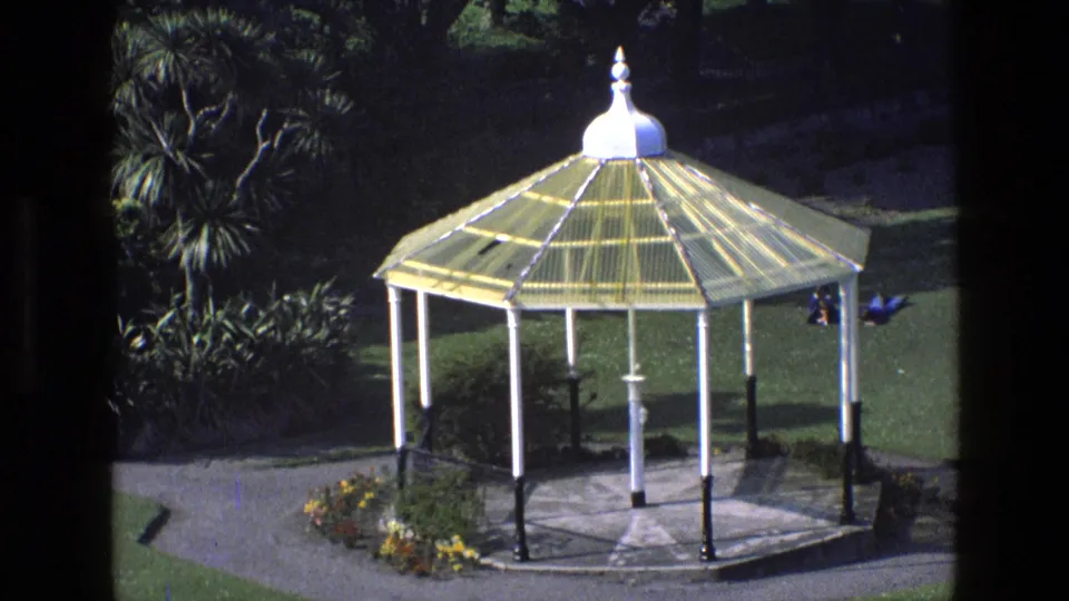 1969 England 8mm frame of a park bandstand with yellow roof panels, white supports and flowerbeds beside the path.