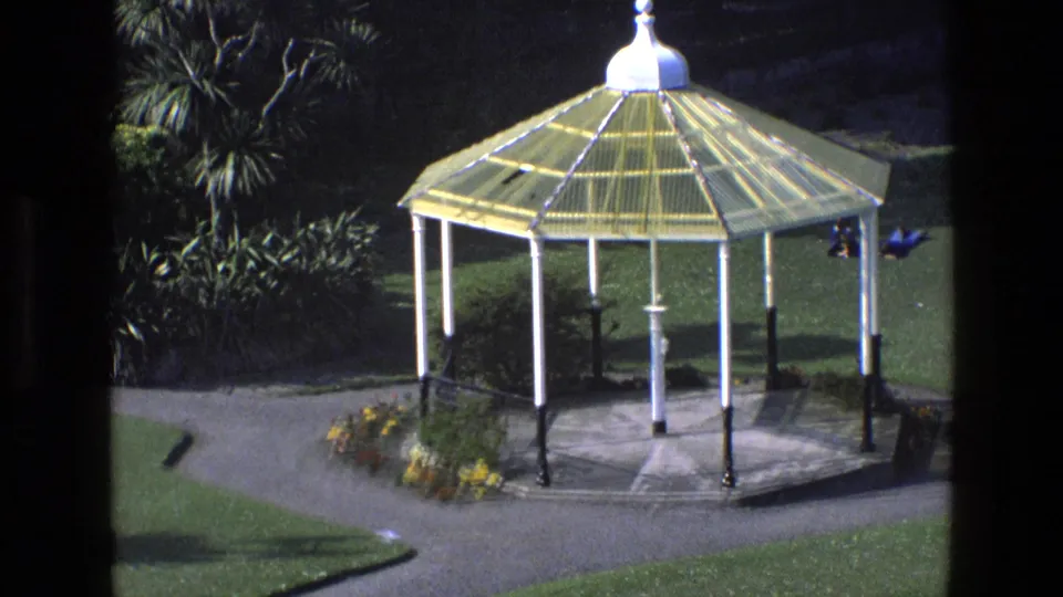 1969 England home-movie view of a yellow-roofed garden bandstand with lawns, trees, paths and small distant visitors nearby.