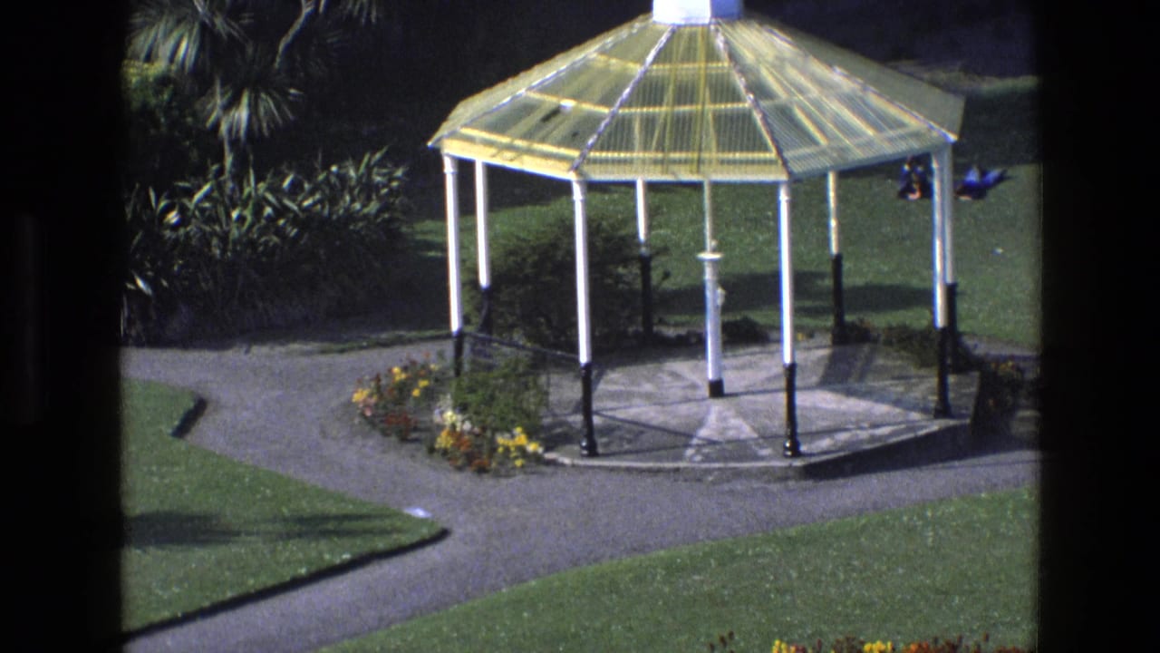 White-and-yellow gazebo beside paths, lawns, and flowerbeds seen through dark framing.