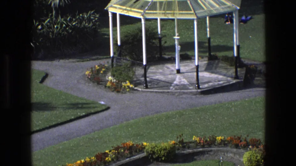 Sunlit 1969 England garden view with an octagonal gazebo, paved walkways, clipped grass, and planted beds.