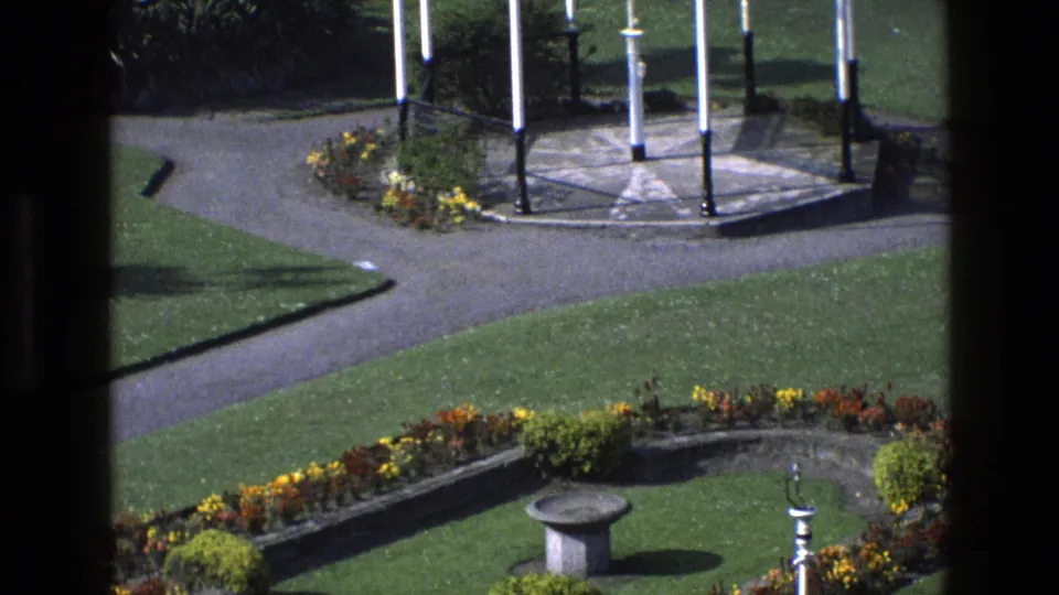 Downward pan in the 1969 England garden reveals flowerbeds, a stone birdbath, lawn, and the lower part of the gazebo.