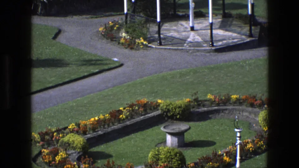 1969 England home-movie frame showing a formal garden bed, stone birdbath, clipped shrubs, paths, and gazebo supports.