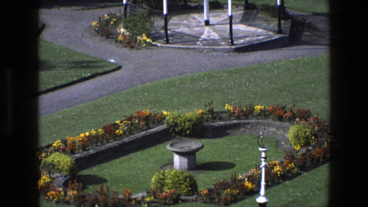 1969 England 8mm frame looking across a formal garden toward paved paths and a white-pillared shelter or bandstand.
