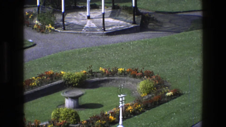 1969 England 8mm frame of a formal garden seen from above, with flowerbeds, paths, lawn, and a stone birdbath.