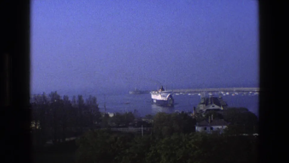 1969 England 8mm frame looking through a dark window frame toward trees, water, a pier, and a distant passenger ship.