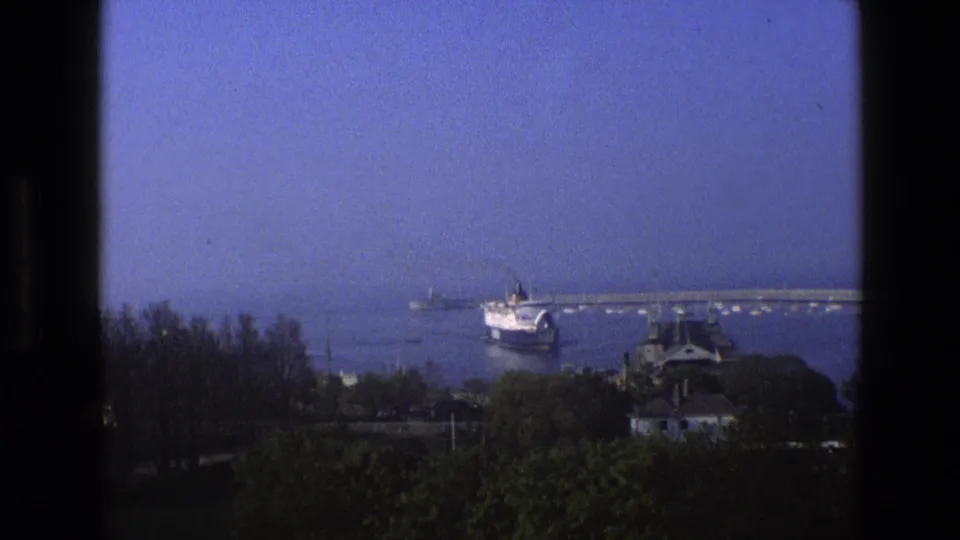1969 England 8mm frame of a harbor view through a dark window frame, with a ship near a pier beyond trees and rooftops.